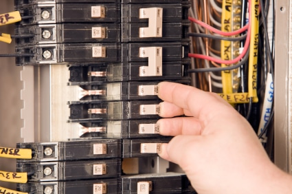 The image shows a hand reaching to flip or adjust a circuit breaker in an electrical panel containing multiple breakers and colorful wiring.
