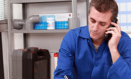 The image shows a technician or service professional in a blue uniform talking on a phone while working in what appears to be a utility room or service area with shelving and equipment in the background.